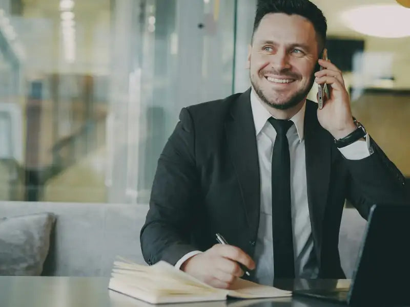 Smiling man taking a phone call while making notes at his desk
